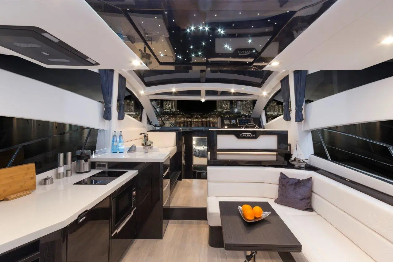 Luxurious yacht interior with modern kitchen, plush seating, oranges on a table, and city lights visible through windows.