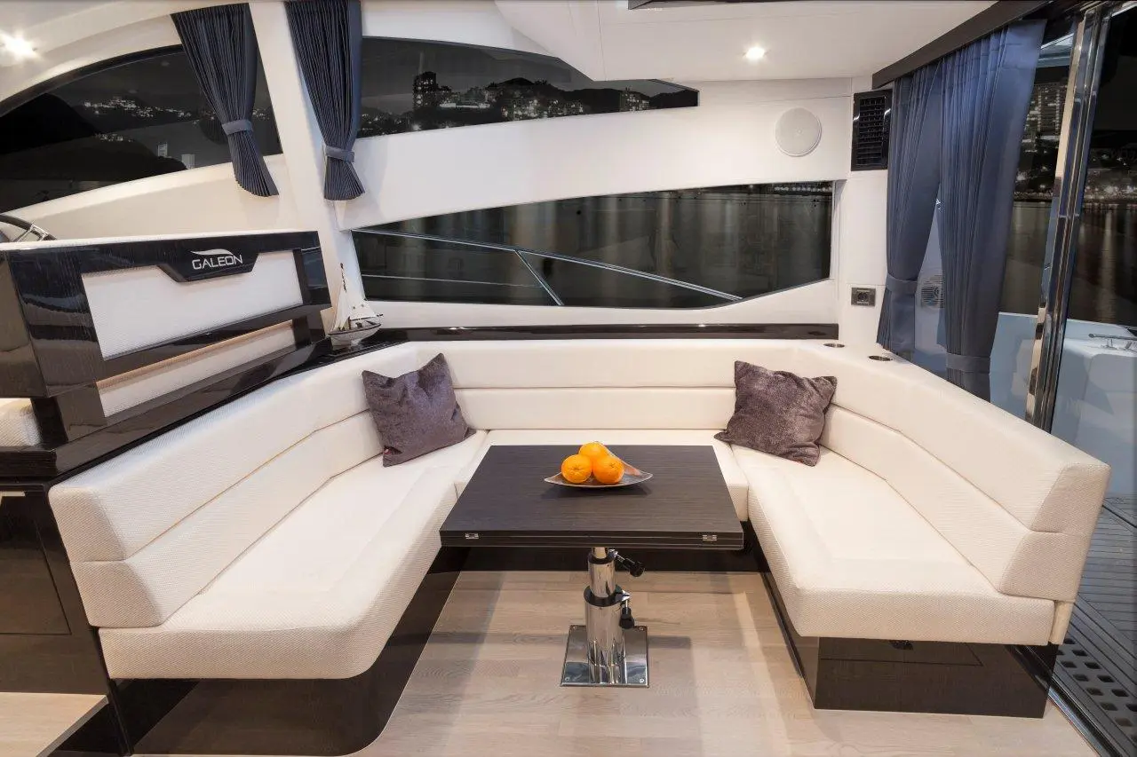Interior of a sleek yacht with a plush seating area, a black table, and a bowl of oranges, overlooking a cityscape at night.