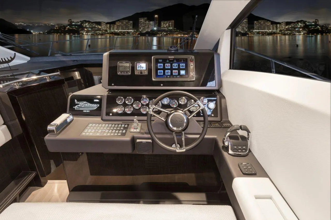 Modern yacht cockpit featuring a steering wheel, digital displays, control panel, and city lights visible through the windows.