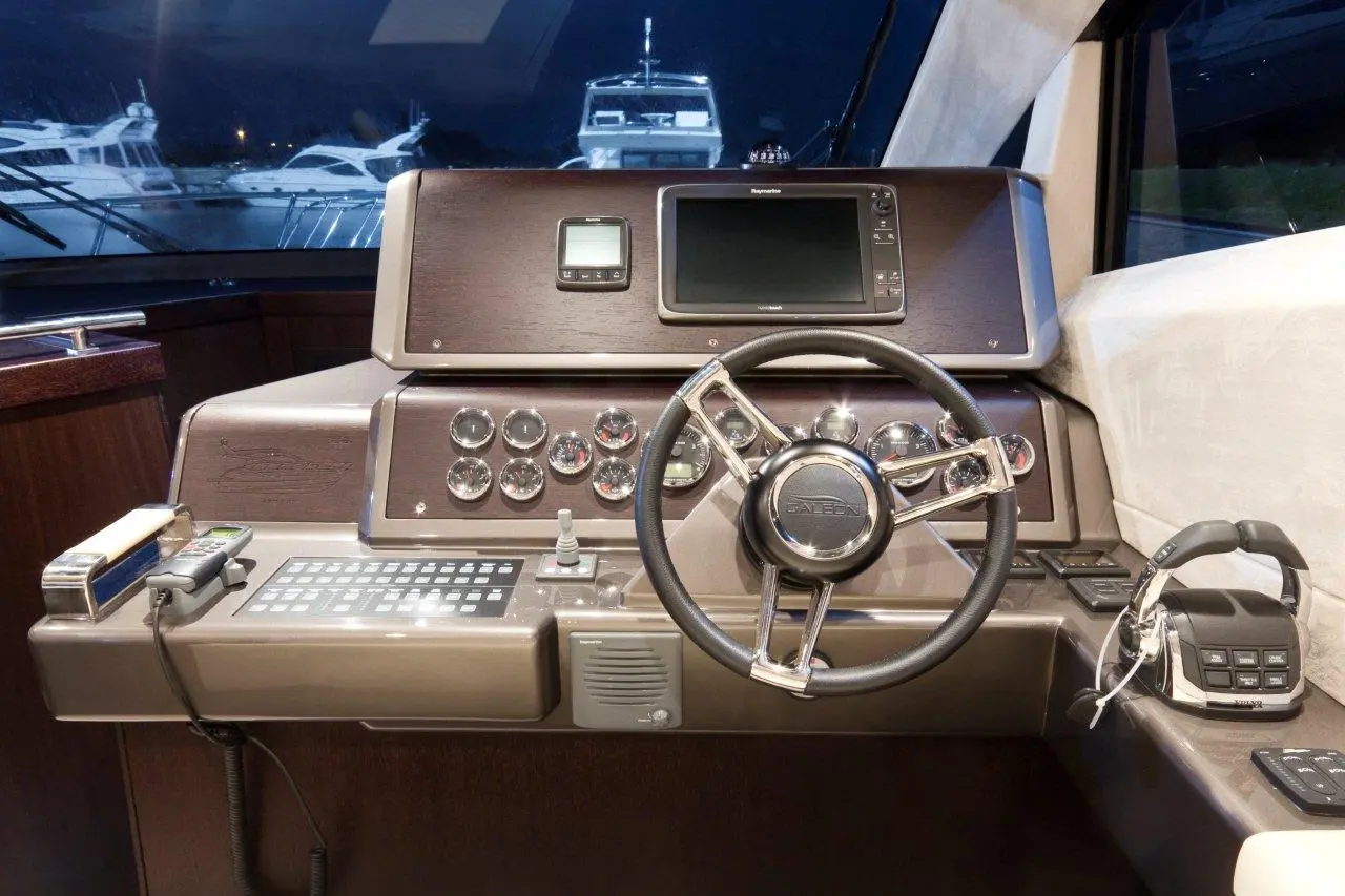 Luxury yacht control panel with a steering wheel, screens, and buttons, viewed at night with boats in the background.