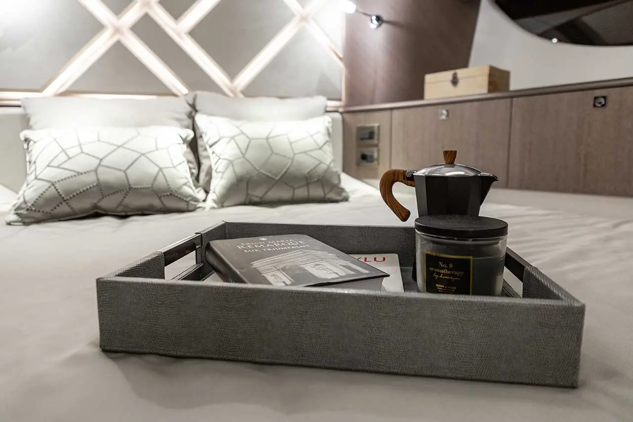 A gray tray on a bed holds a coffee maker, a candle, and a book, with stylish pillows and decorative wall lighting behind.
