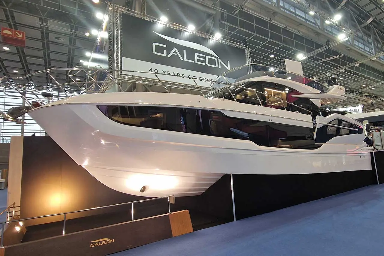Galeon yacht displayed at a boat show, featuring a sleek white hull and large windows, with a large sign reading 