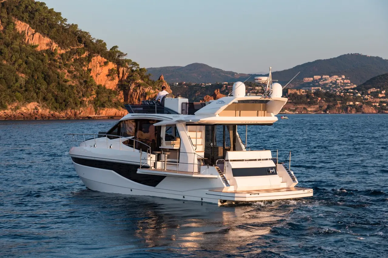 A luxury white motorboat navigates calm waters near rocky coastline under a clear sky, with mountains and buildings in the background.