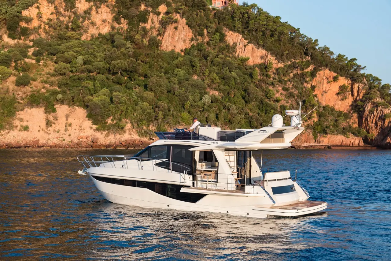 Luxury yacht cruising on water near a rocky coastline, with a person enjoying the view from the upper deck.