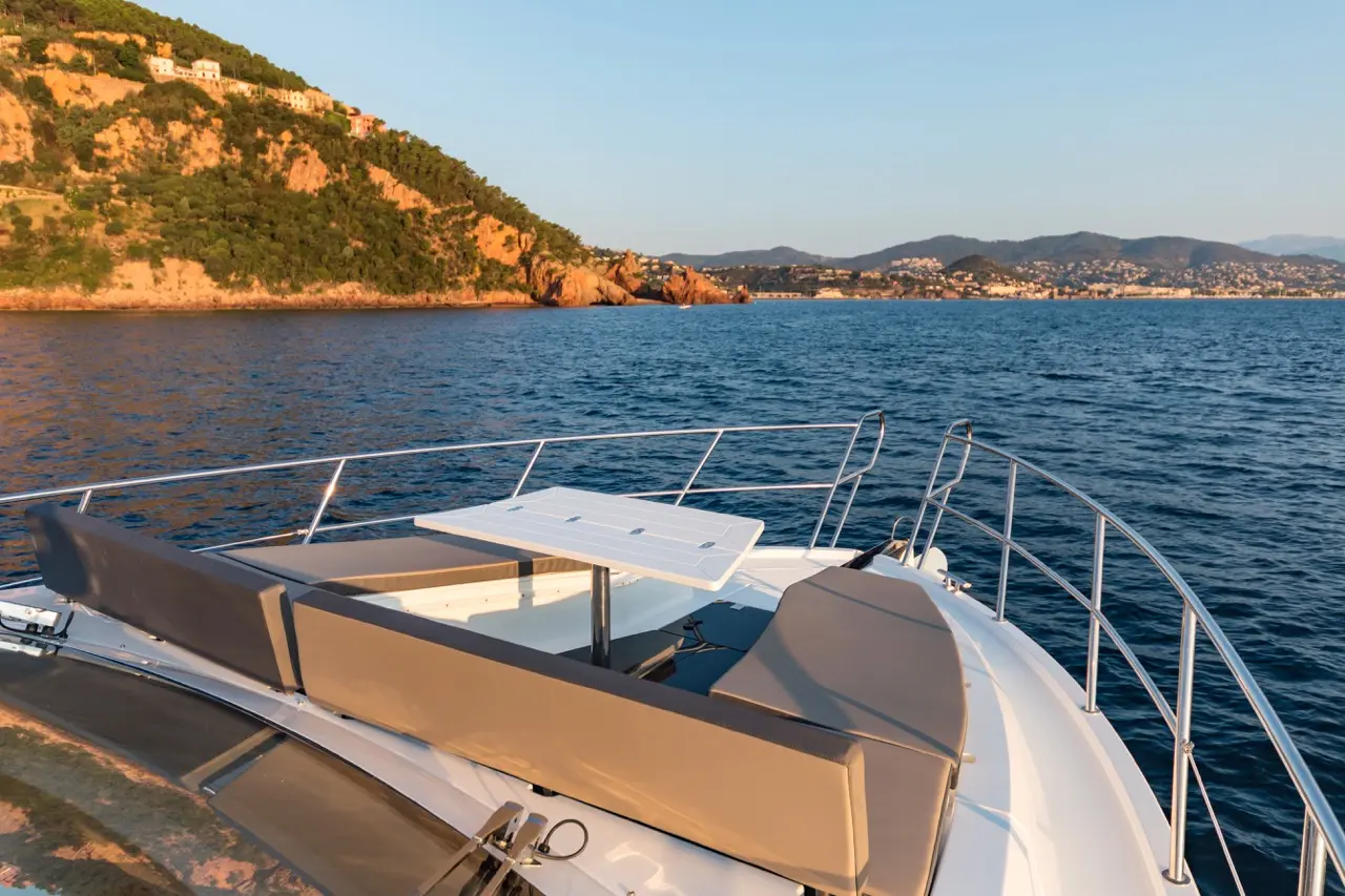 Luxury yacht deck with cushioned seating and a table, overlooking calm waters and a rocky coastline at sunset.