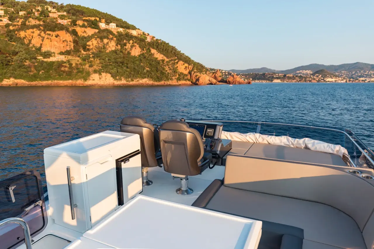 Yacht interior with two gray captain chairs, controls, and a cooler, overlooking calm waters and hills at sunset.