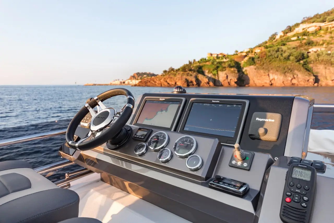 Boat cockpit view showcasing a steering wheel, navigation screens, and control instruments with a coastal backdrop.