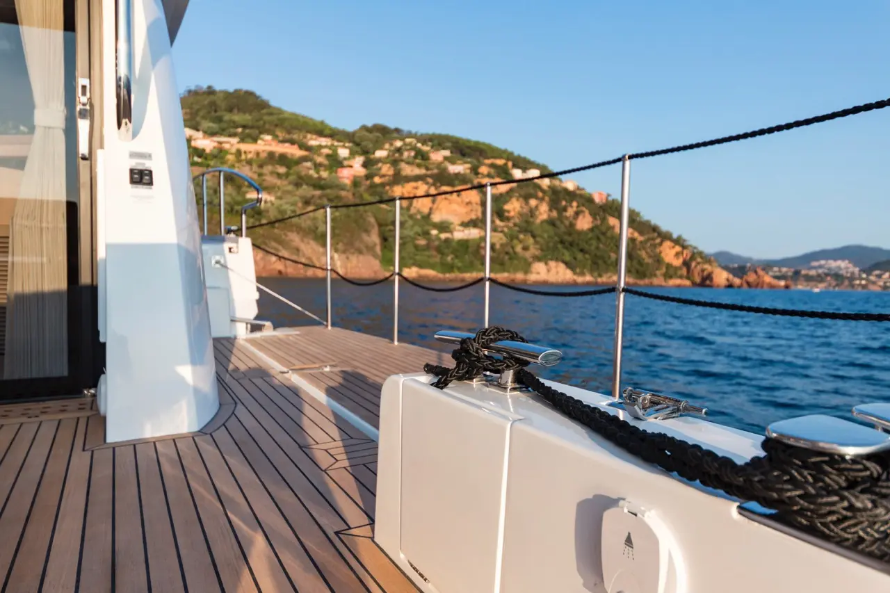 Deck of a yacht with wooden flooring, mooring cleat, and a view of a coastal landscape under a clear blue sky.
