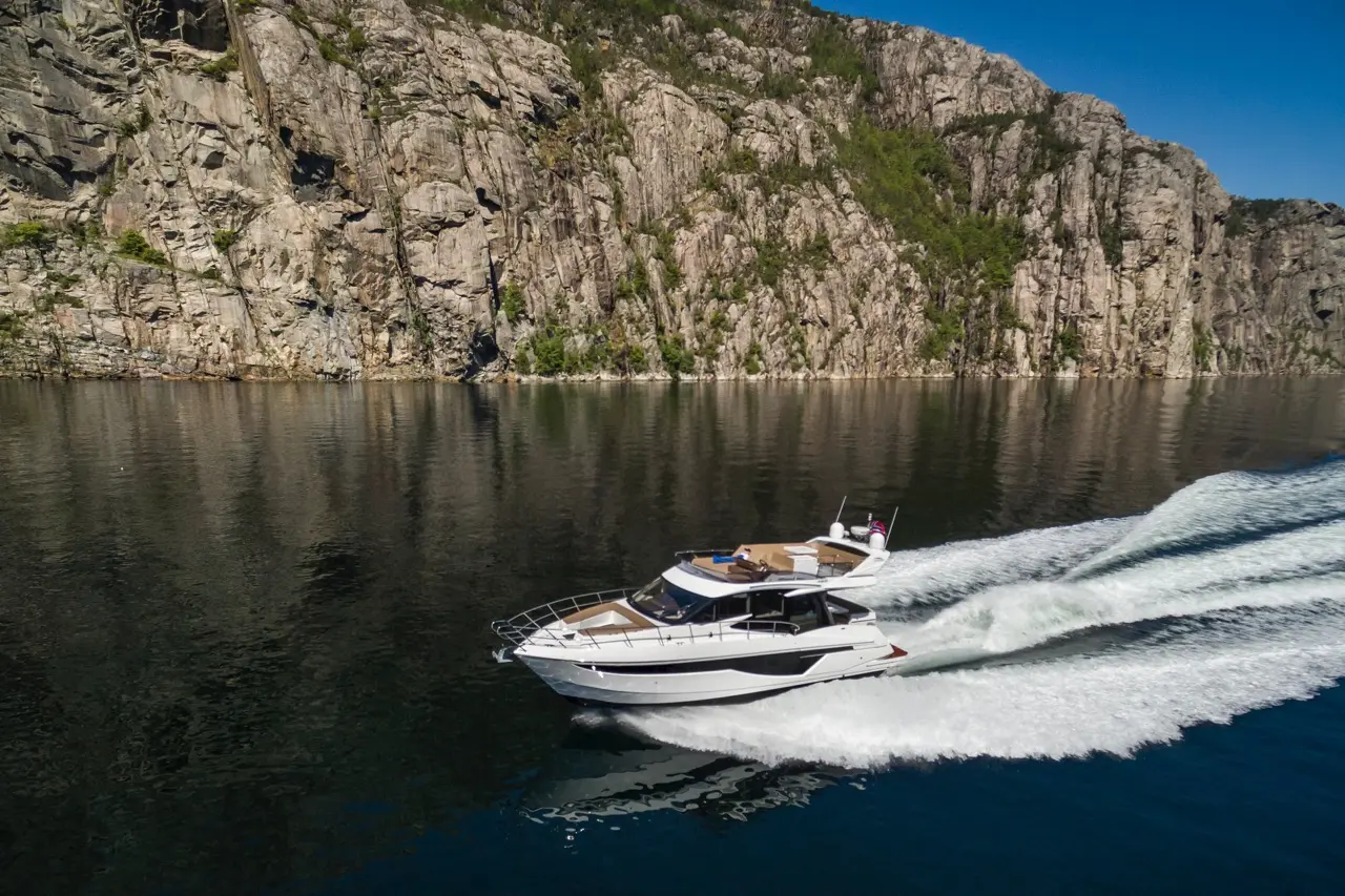 A white motor yacht speeds across calm waters, with rocky cliffs and greenery reflecting in the water's surface.