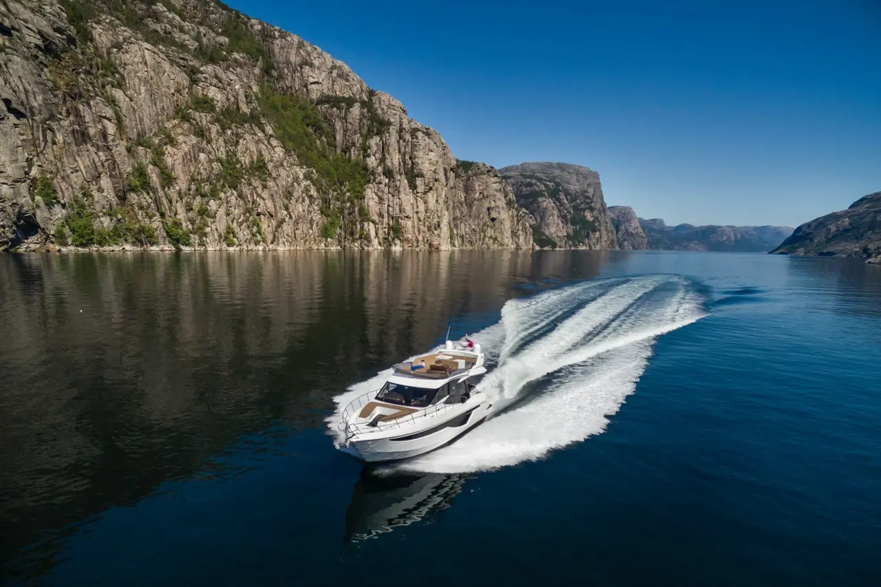 A sleek motorboat speeds through calm water, surrounded by towering rocky cliffs under a clear blue sky.