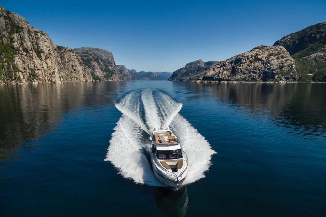 A sleek motorboat cuts through calm waters, leaving a wake against rocky cliffs under a clear blue sky.