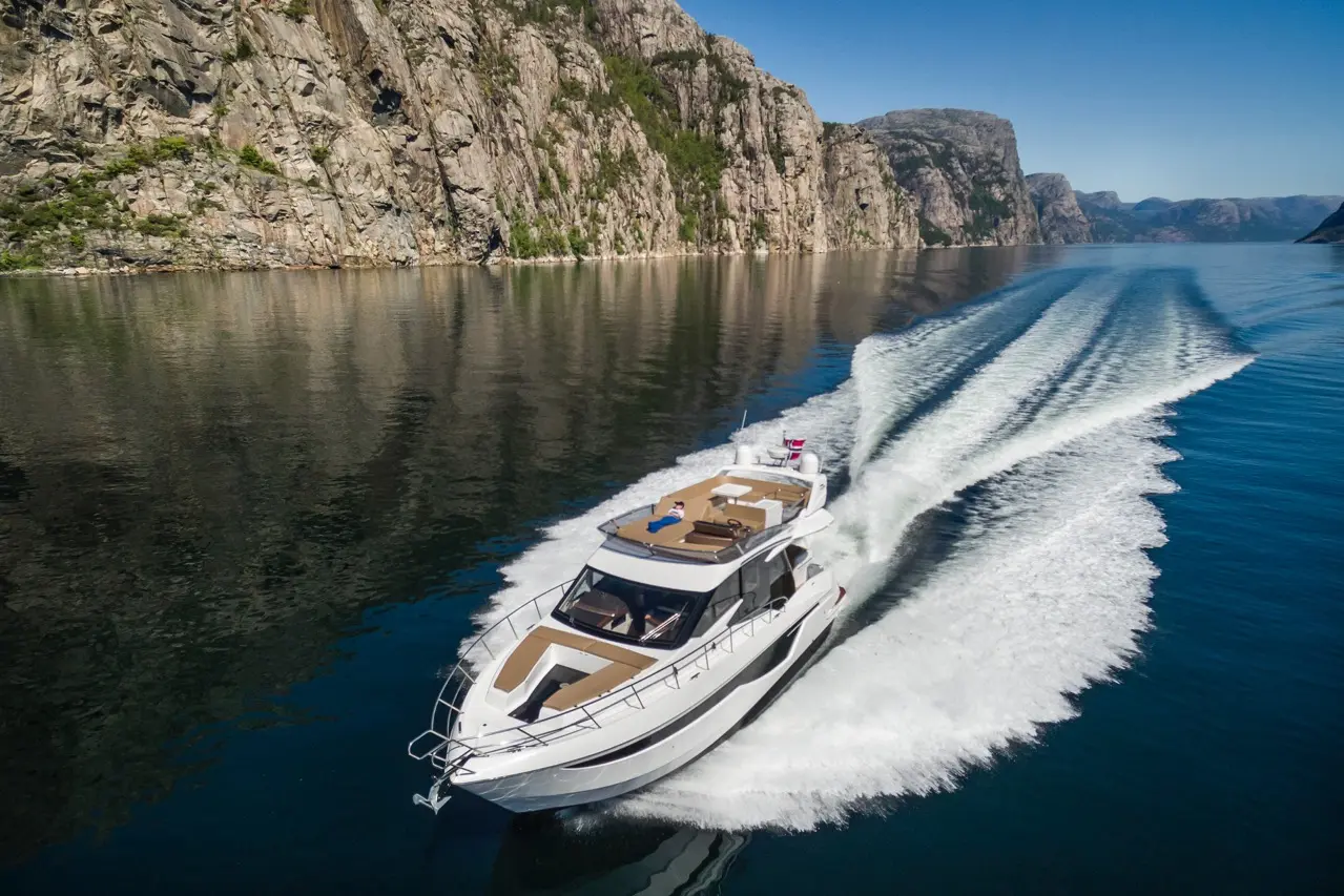 A motorboat cruises through a calm fjord surrounded by rocky cliffs, leaving a wake on the water's surface.