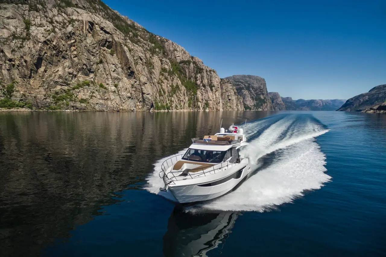 A white motorboat navigates a calm fjord, surrounded by steep rocky cliffs under a clear blue sky.