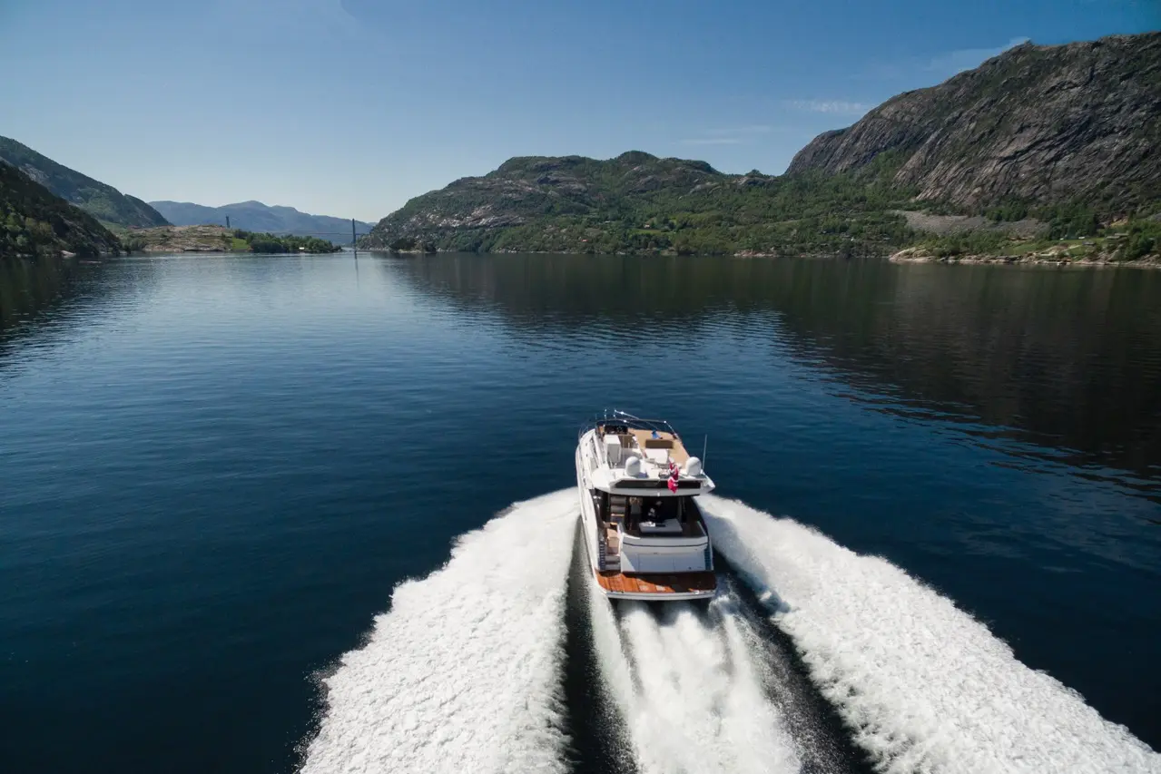 A motorboat speeds across calm waters, leaving a white wake, surrounded by green mountains under a clear blue sky.