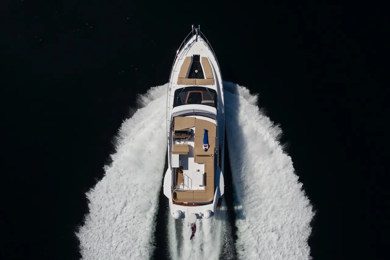 Aerial view of a white yacht speeding on dark water, leaving a wake, with a person being towed on a wakeboard.