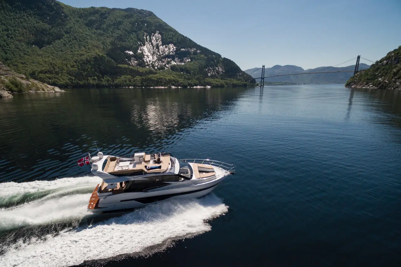 Yacht cruising on calm waters with a scenic backdrop of mountains and a bridge under a clear blue sky.