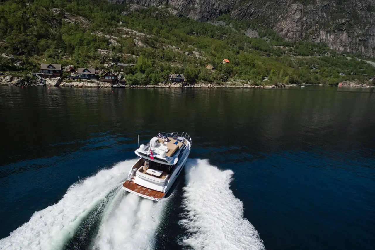 A motorboat leaves a wake in a calm fjord, surrounded by green hills and wooden houses under a clear sky.