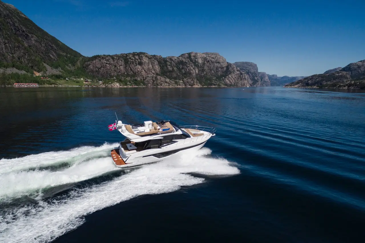 A luxury yacht with a British flag cruises through calm waters, surrounded by dramatic rocky mountains and clear blue skies.