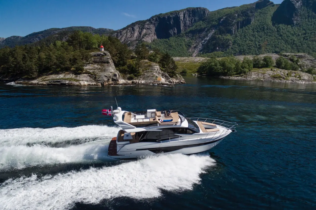 A luxury yacht speeds through calm waters, with rocky hills and a lighthouse in the background under a clear blue sky.