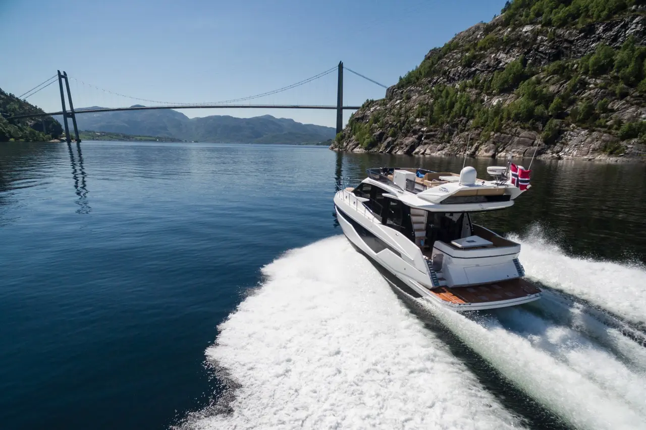 A luxury yacht with a Norwegian flag glides across calm waters, with a suspension bridge and green hills in the background.