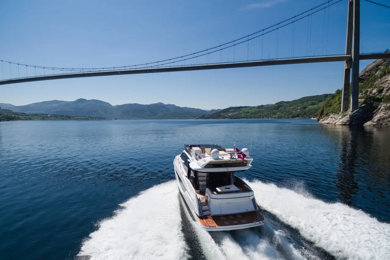 A luxury yacht cruising on calm waters beneath a large bridge surrounded by green hills and clear blue skies.