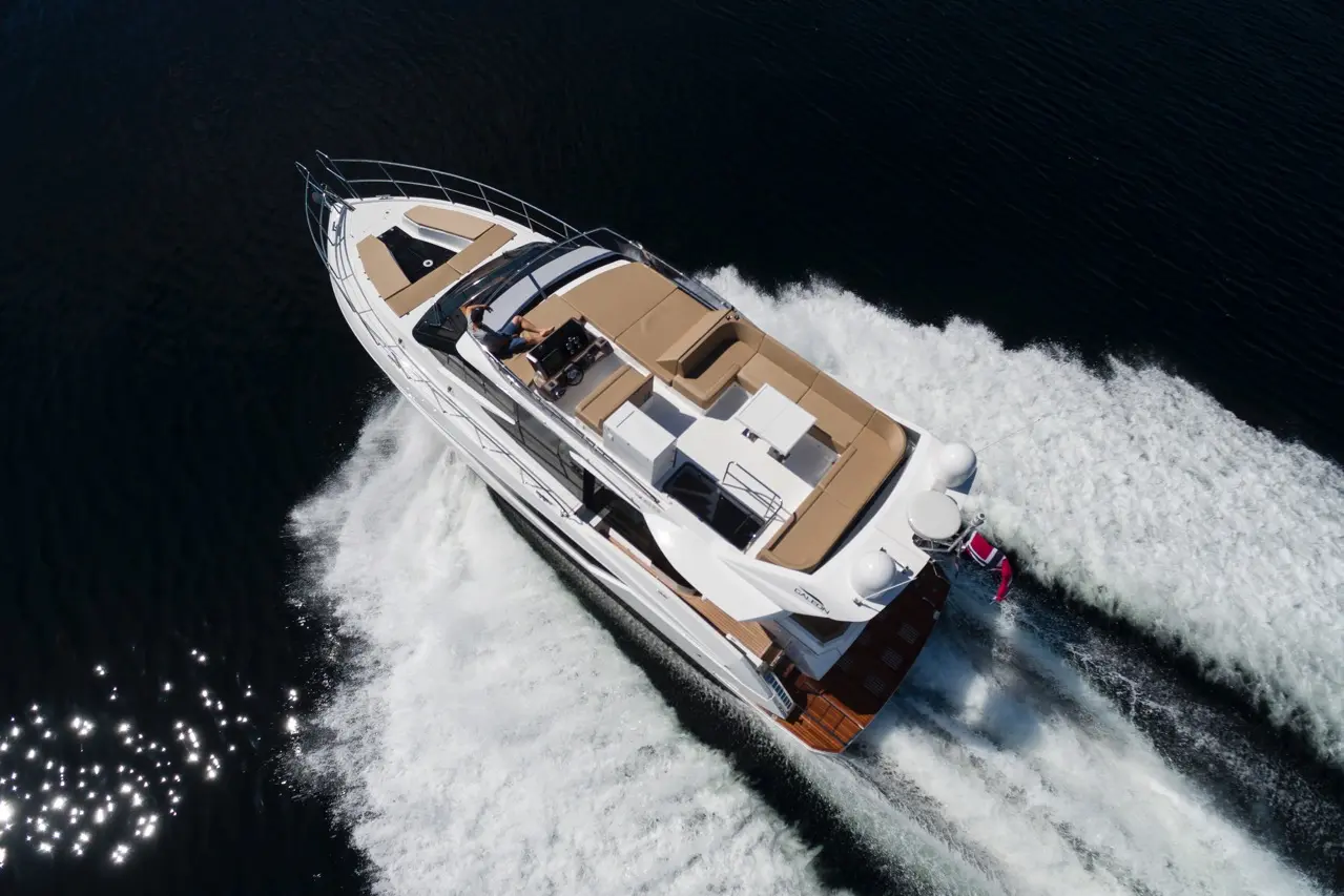 Luxury motor yacht cruising on calm waters, creating a white wake, with a person at the helm enjoying the sun.