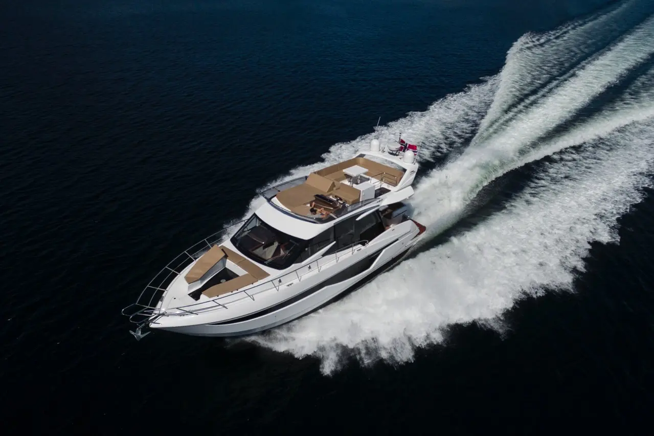 Luxury motor yacht gliding over deep blue water, leaving a white wake; deck features sun loungers and a small table.