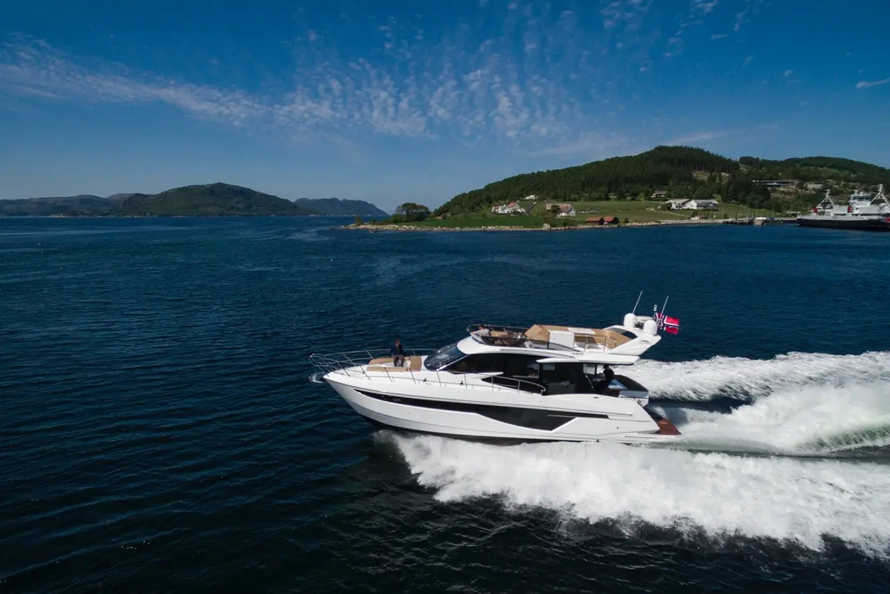 A white luxury yacht glides through blue waters near a green hill, with small houses and a Norwegian flag visible.