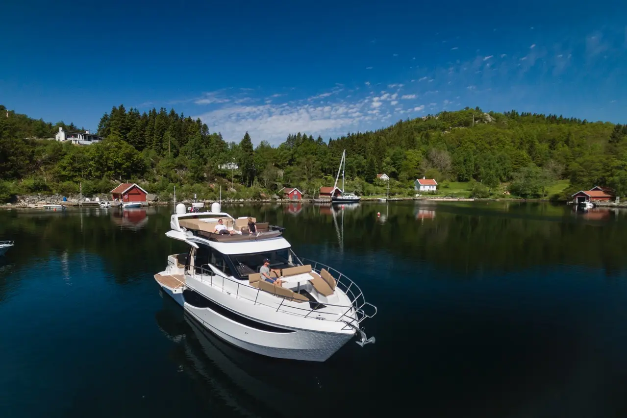 A luxury yacht anchored in a serene bay, surrounded by green hills and rustic red cabins under a clear blue sky.