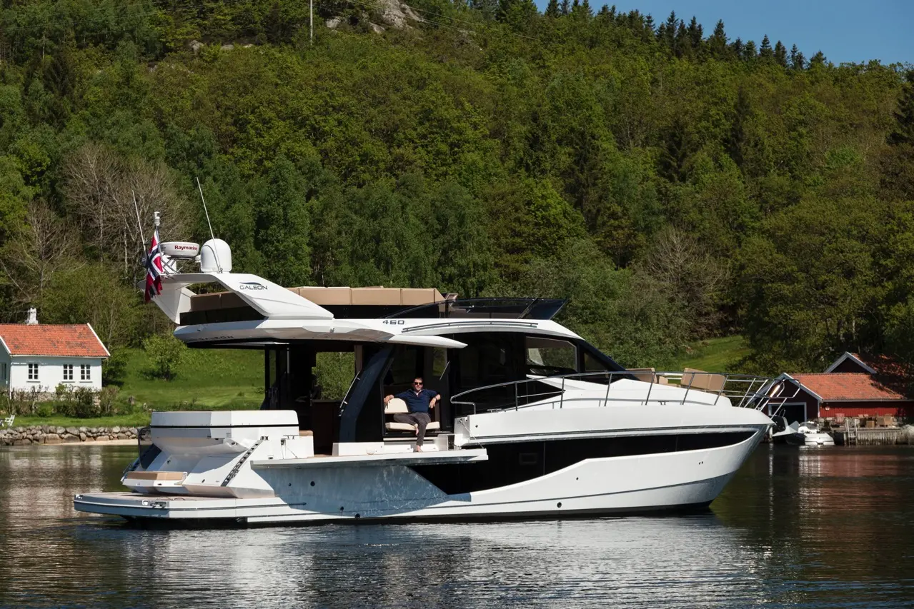 Luxury Caleón 460 yacht docked on calm water, with a man relaxed on deck, surrounded by lush greenery and a quaint house nearby.
