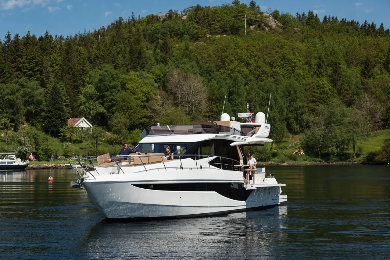 A luxury motorboat with people on deck navigating a calm waterway, surrounded by lush green hills and a house on shore.