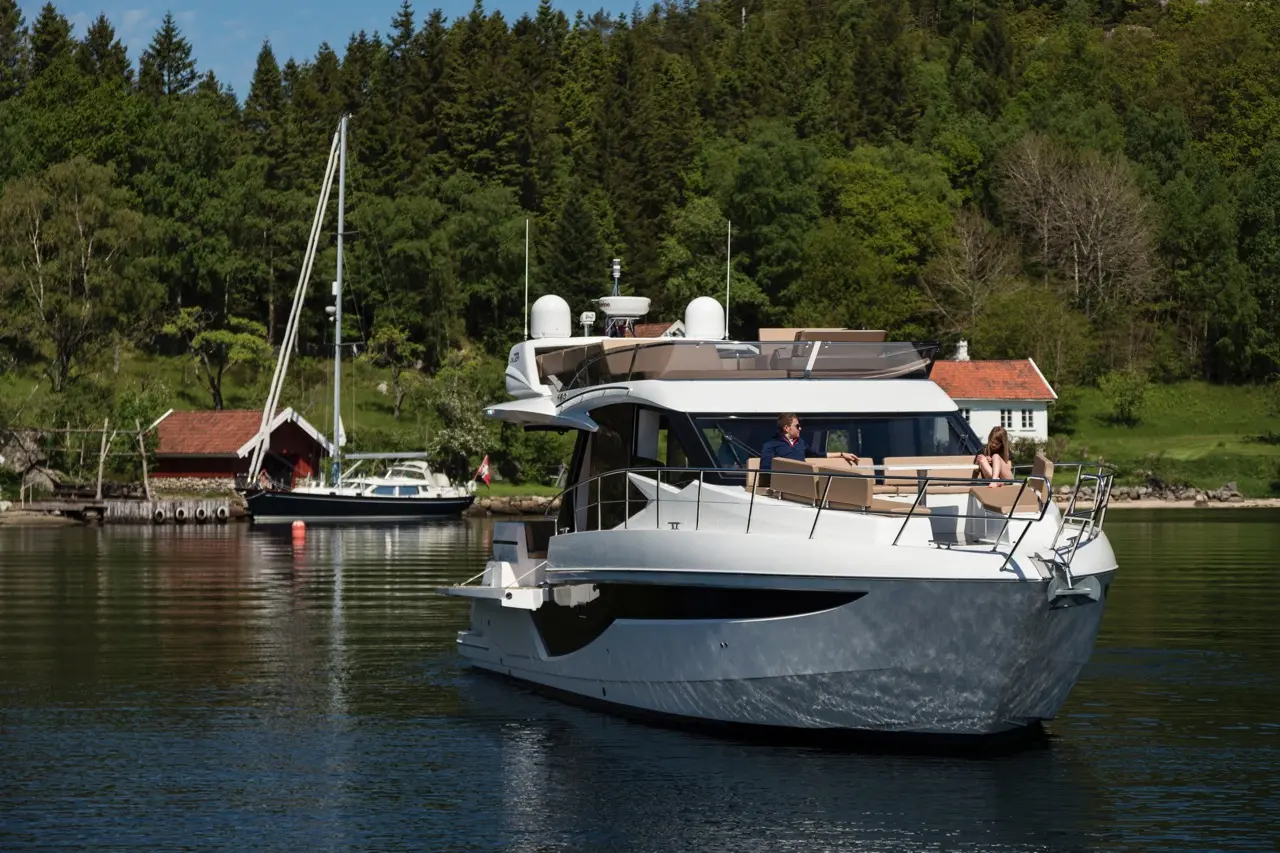 A luxury yacht with two people on the deck, moored near a sailboat and a red boathouse, surrounded by lush greenery.