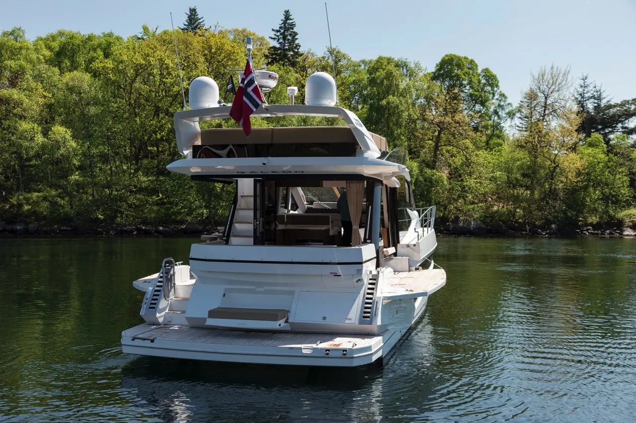 Luxury yacht docked in calm water, featuring a Norwegian flag and a backdrop of lush green trees under clear skies.