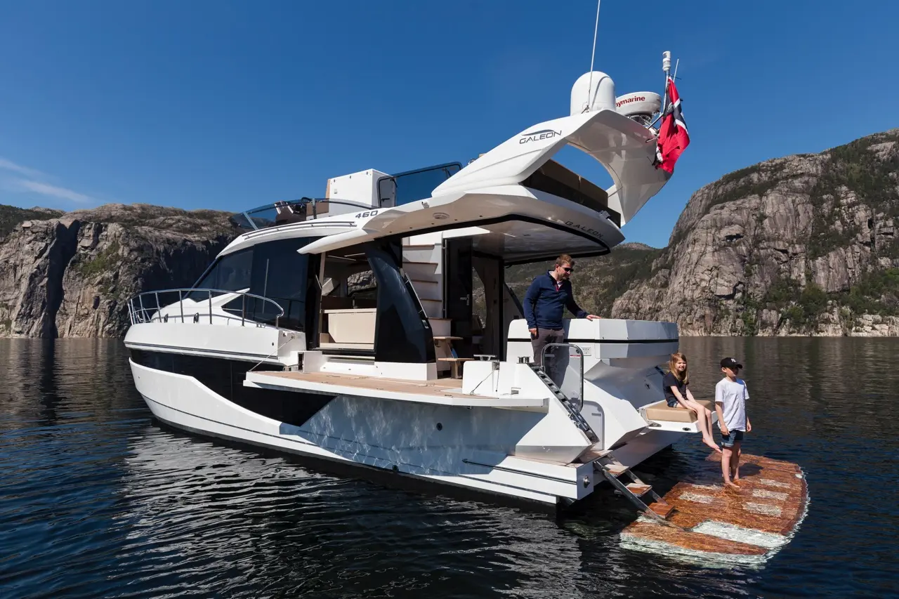A sleek white yacht is anchored, with two children playing on a floating platform while an adult supervises.