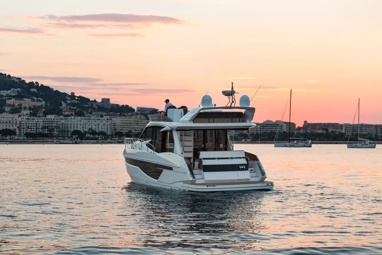 Luxury motorboat cruising in calm waters at sunset, with a silhouette of a man on board and coastal buildings in the background.