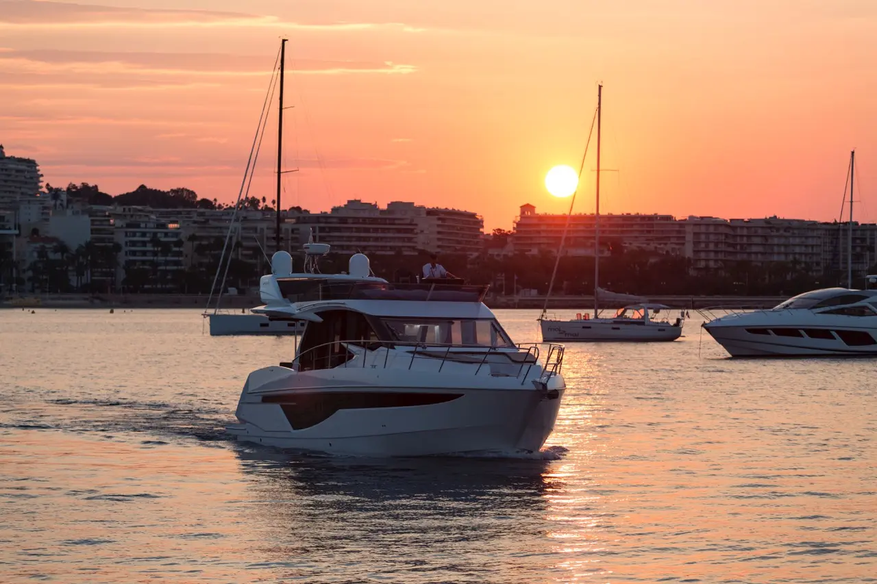 A white yacht sails on calm waters at sunset, with a vibrant orange sky and silhouetted boats in the background.