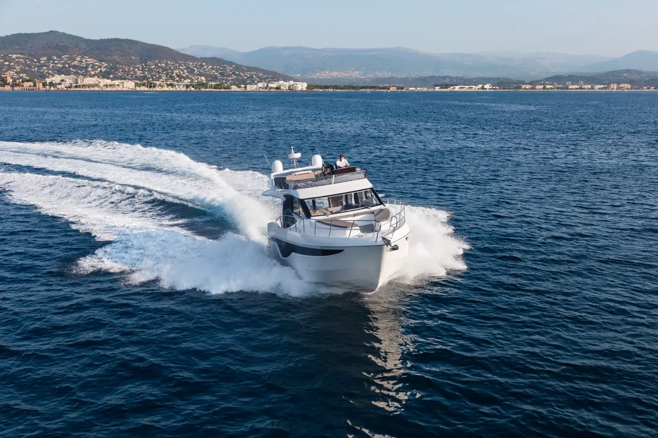 A modern motorboat speeds across a calm blue sea, creating white foam trails against a coastal backdrop.