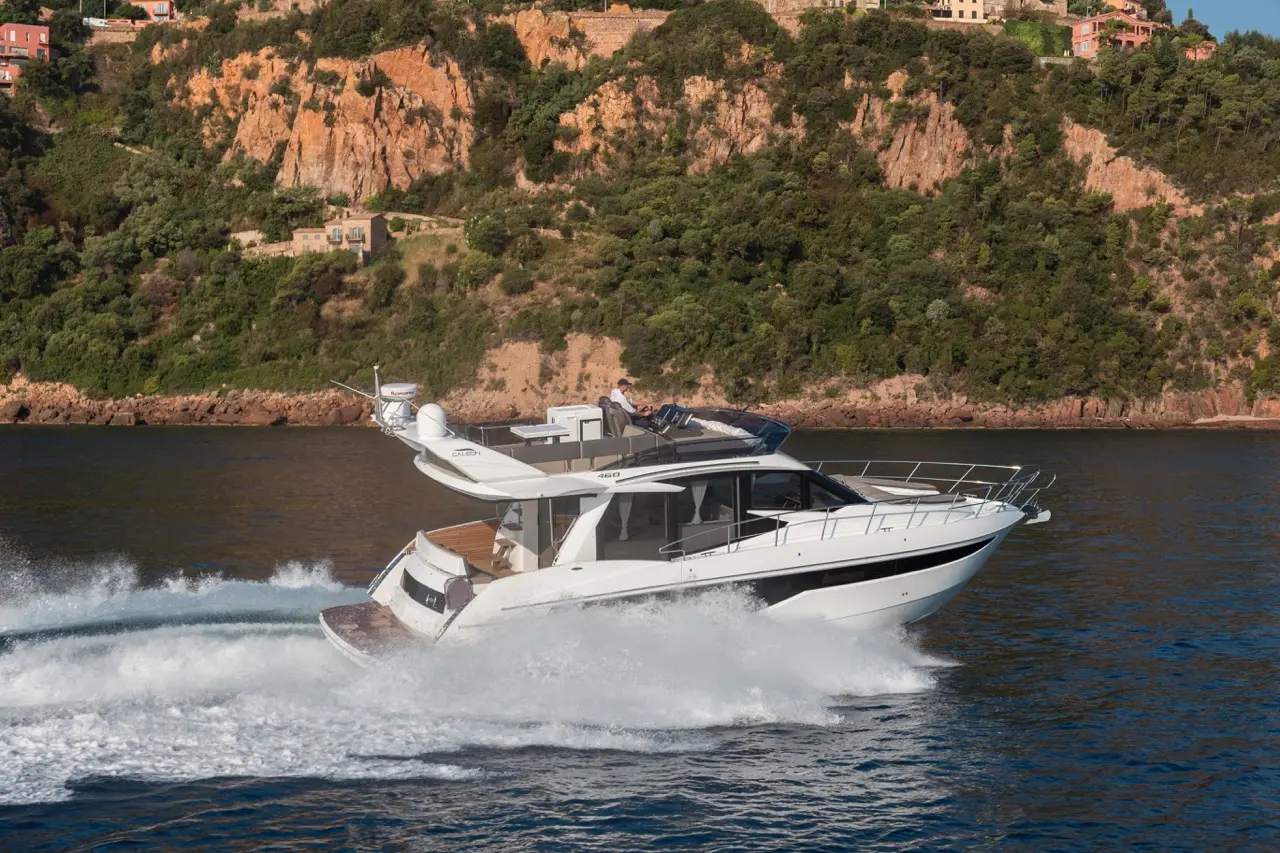 A luxury yacht speeds through blue waters, with a rugged cliffside and greenery in the background, showcasing scenic coastal beauty.