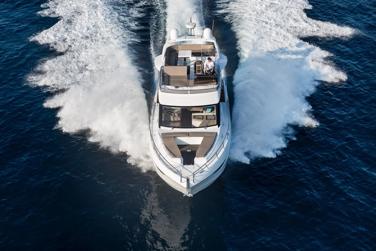 Luxury motor yacht gliding over blue water, creating white foam trails; features a sun deck and a seated captain navigating.