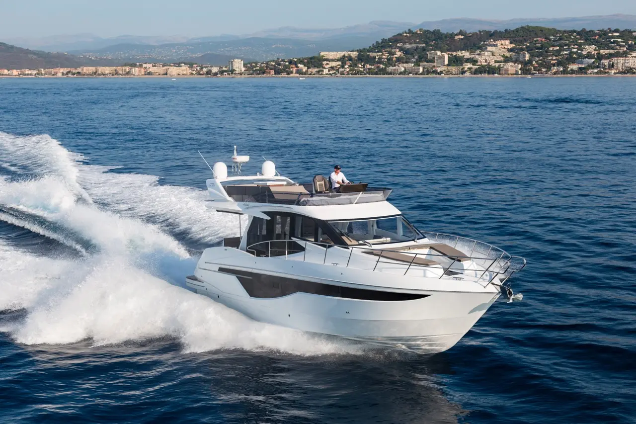 A luxurious yacht speeds across the calm sea, with a man at the helm and a coastal city visible in the background.