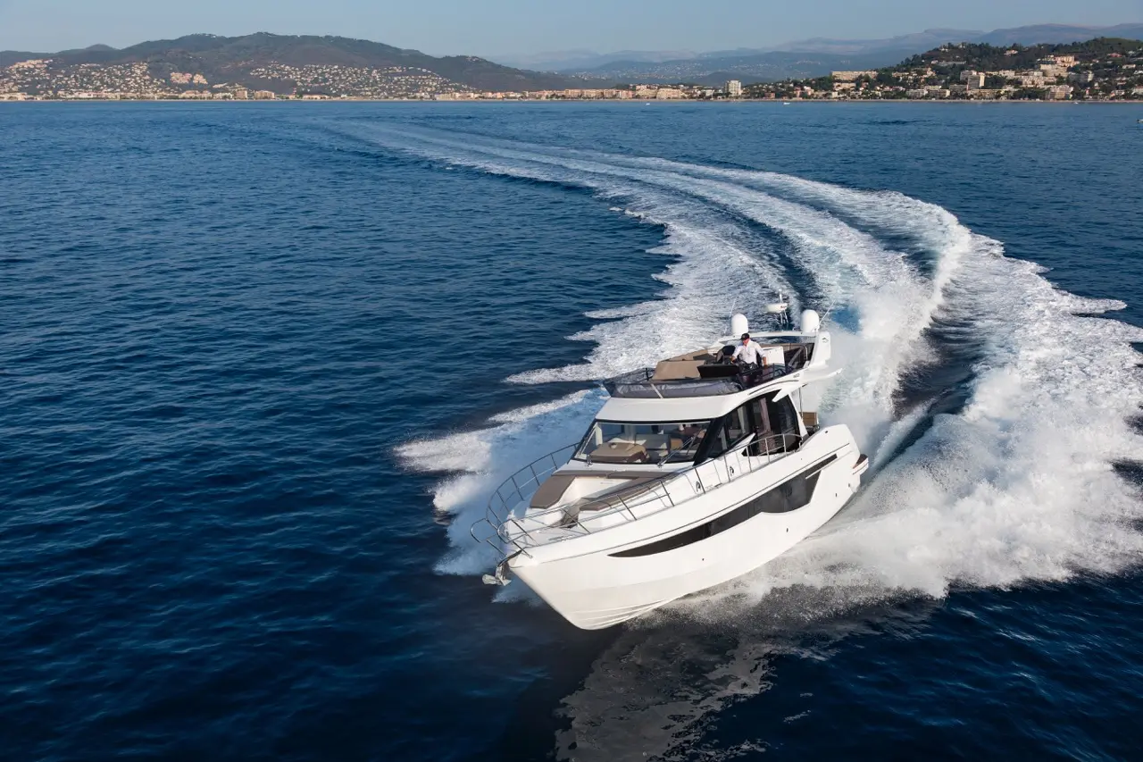 Luxury yacht cruising on blue waters, leaving a white wake, with hills and buildings in the background under clear skies.