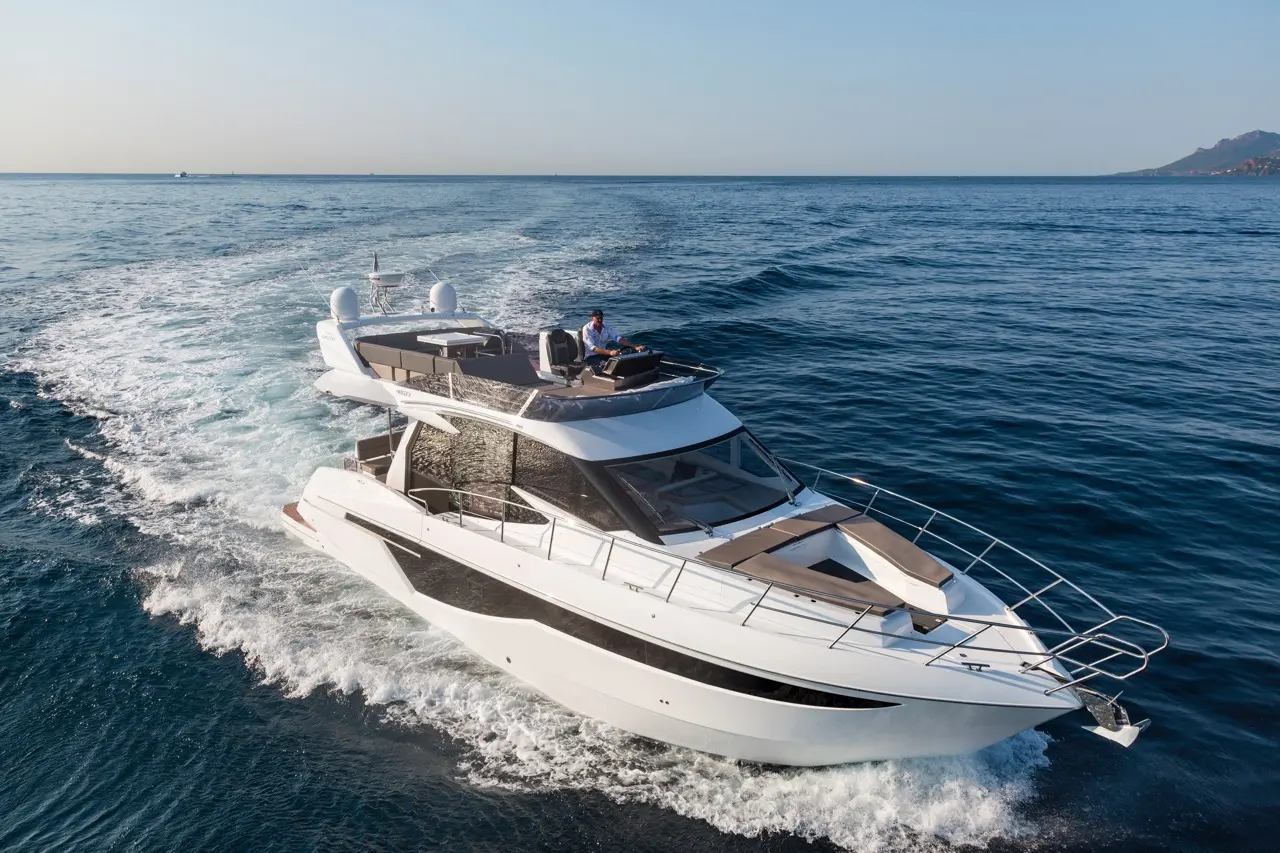 A luxury motor yacht cruises through clear blue water, with a man steering and an elegant design prominent.