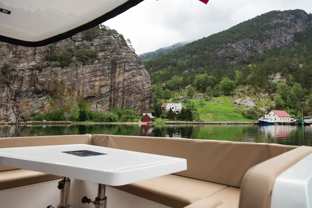 View from a boat showing a white table, steep cliffs, green hills, and houses by the water under a cloudy sky.