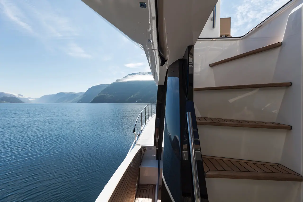 View from a yacht showing stairs leading up to the deck, with serene water and mountains in the background under a clear sky.
