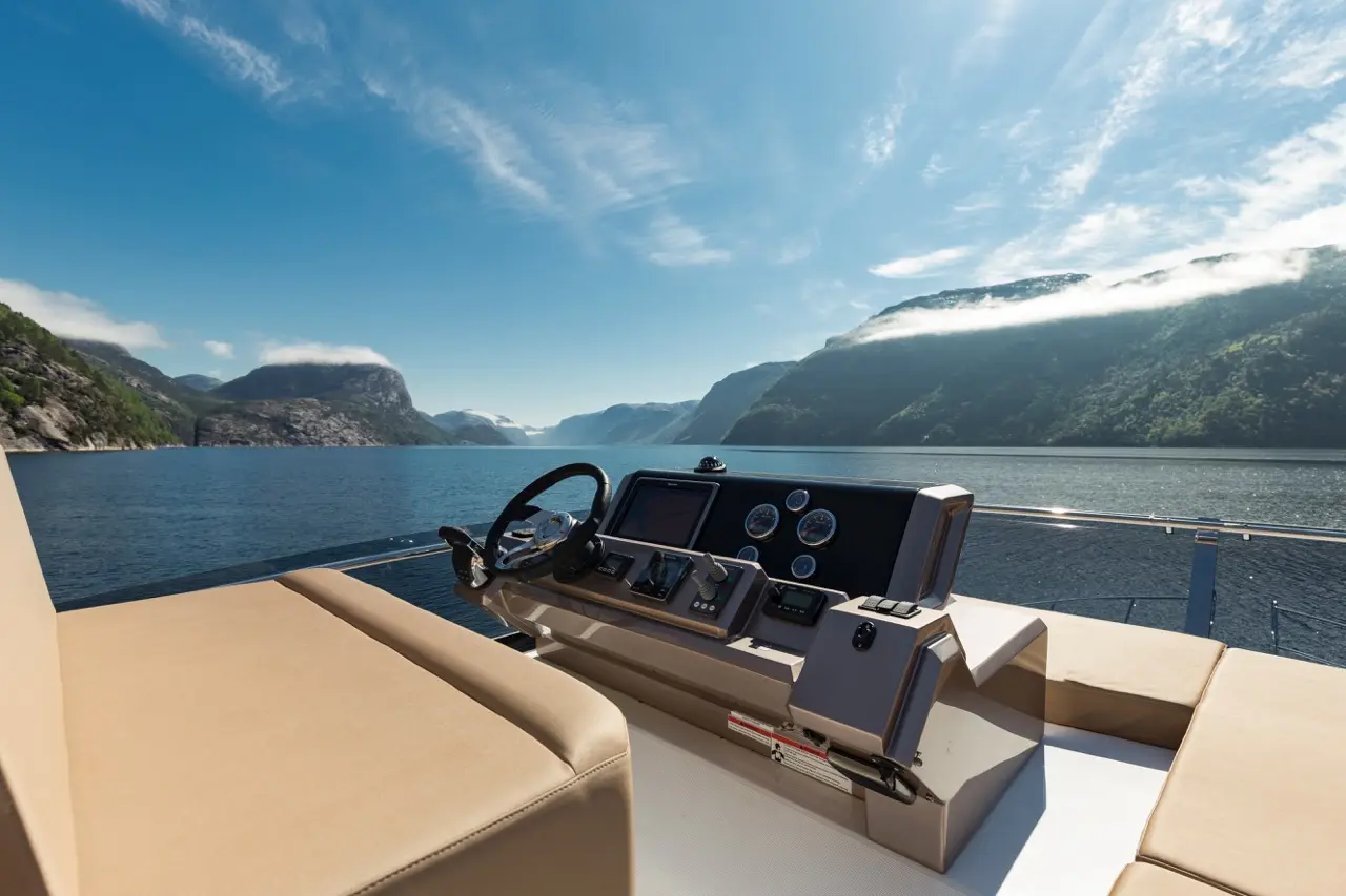 Steering wheel and dashboard of a yacht with scenic fjord landscape and clear blue sky in the background.