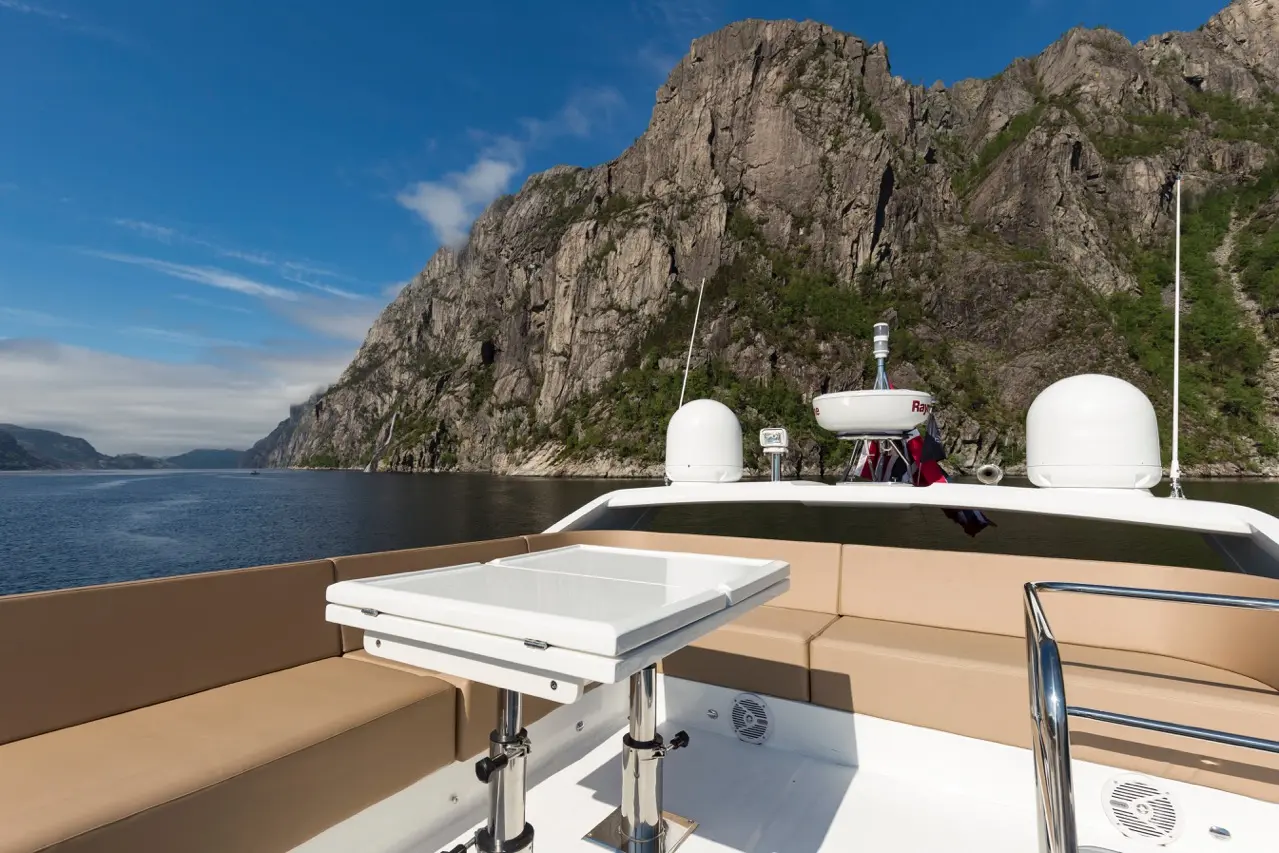 A scenic view from a boat featuring mountains, a calm waterway, and a white table on the boat's deck under a blue sky.