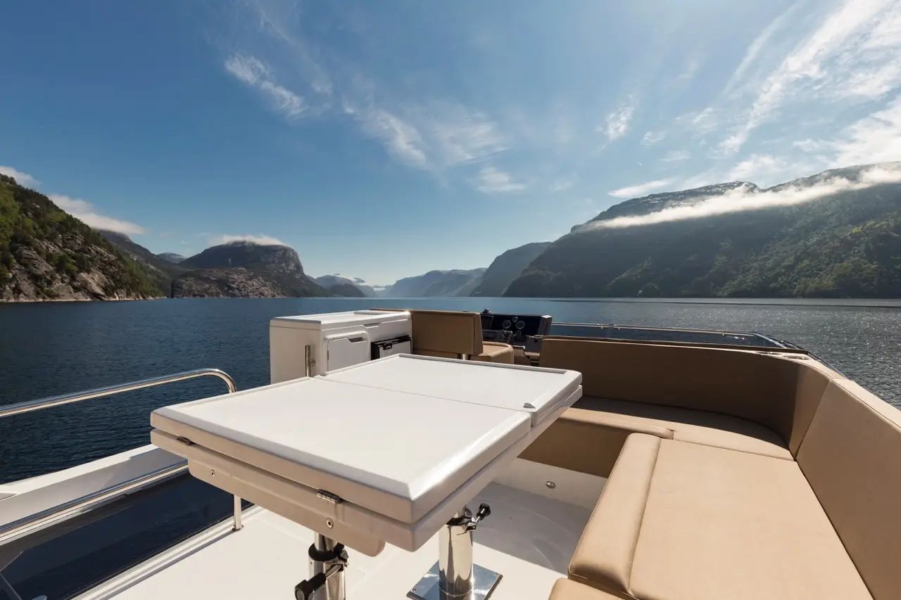 Luxury yacht deck with a foldable table, cushioned seating, and scenic fjord view under a clear blue sky.