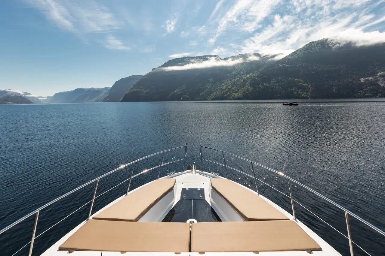 View from the bow of a yacht overlooking a calm fjord with mountains and clouds in the background.