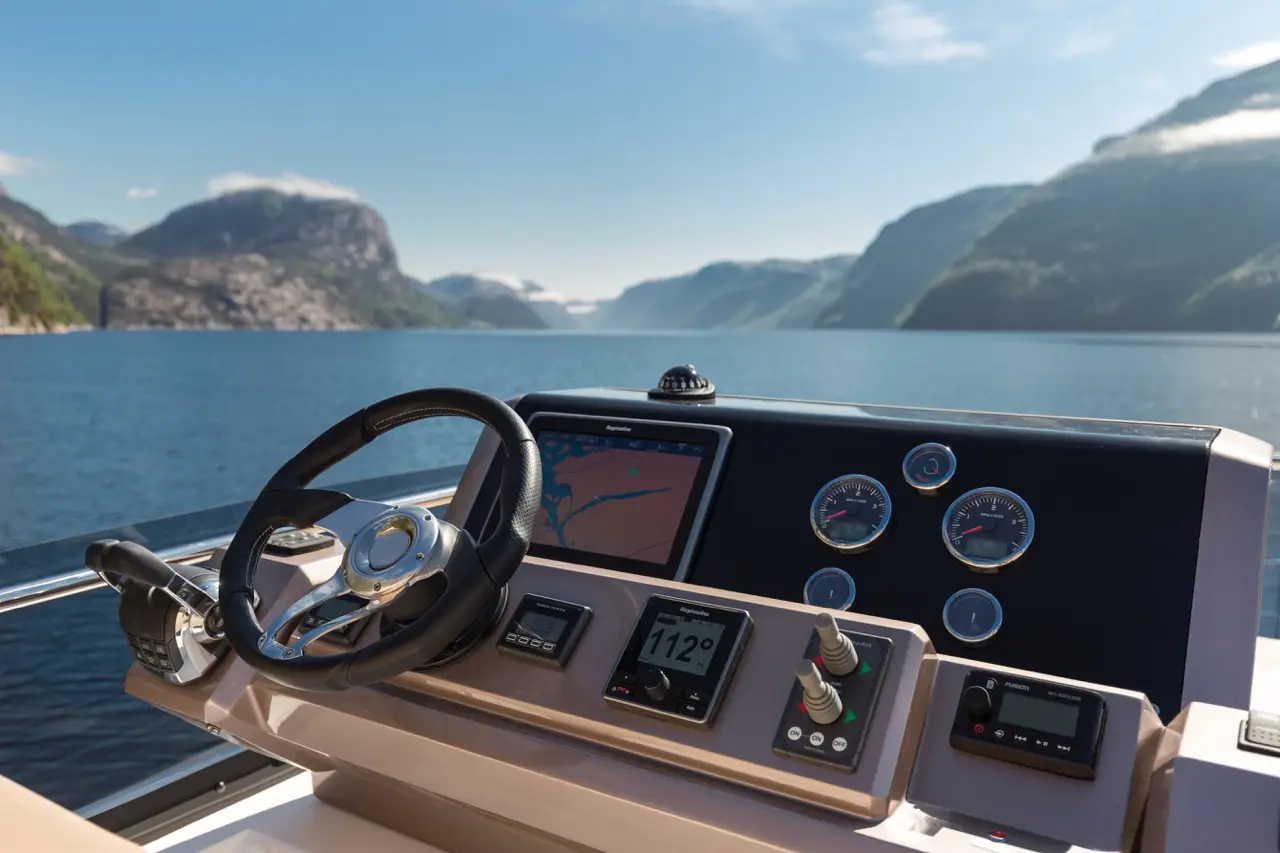 Boat dashboard with steering wheel, navigation screen showing a map, and gauges against a backdrop of mountains and water.