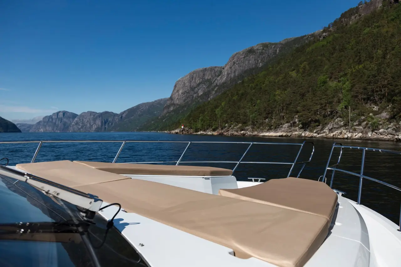A luxury boat deck with beige cushions overlooks a serene waterway surrounded by mountains and trees under a clear blue sky.